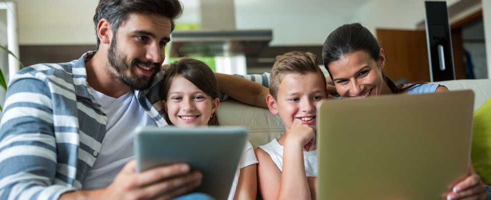 Minacce Informatiche 980x400 Famiglia sorridente seduta sul divano mentre guarda insieme uno schermo di laptop; il padre tiene anche un tablet